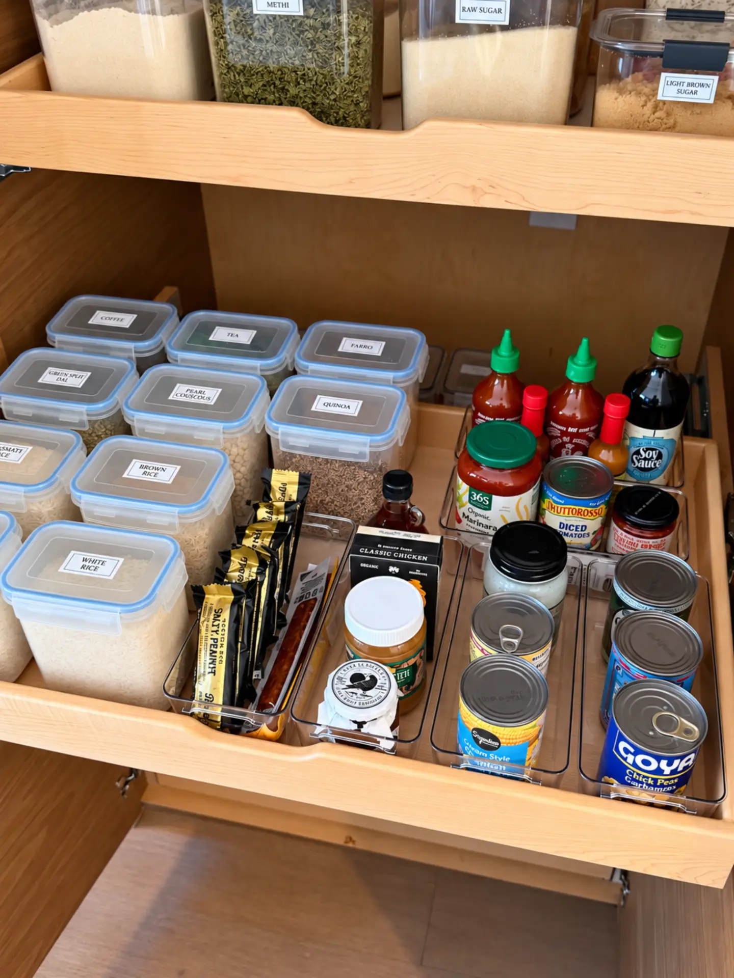 Pull-out pantry drawer with labeled clear containers of rice and grains beside acrylic bins of snack bars, condiments, and canned goods