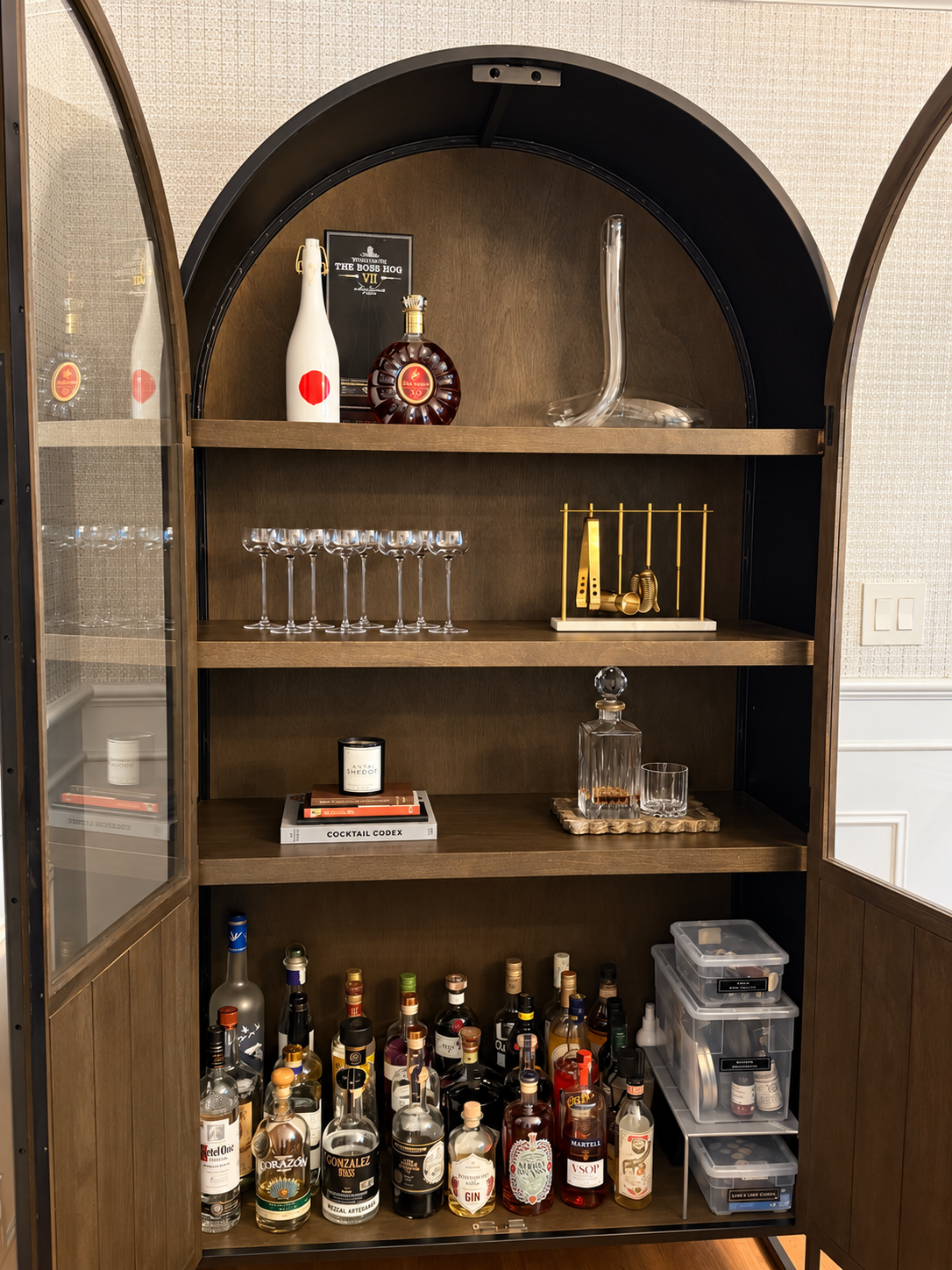 Styled arched bar cabinet with glass doors open, a row of cocktail glasses and a gold bar tool set on the upper shelves, a candle resting on books beside a decanter on a gold tray, and liquor bottles with clear bins below