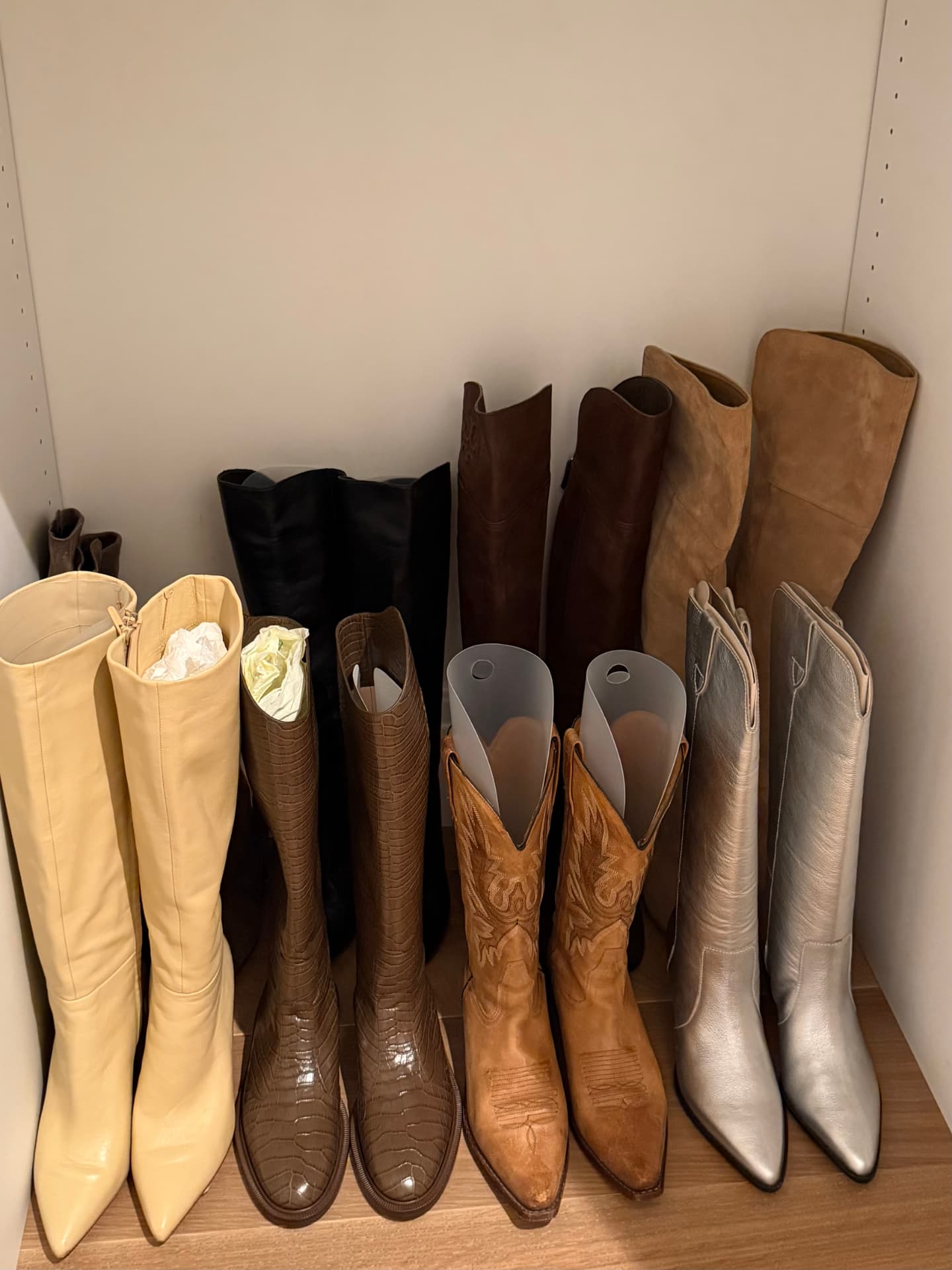 Row of tall boots in cream, brown, tan, and silver lined up on a wooden shelf
