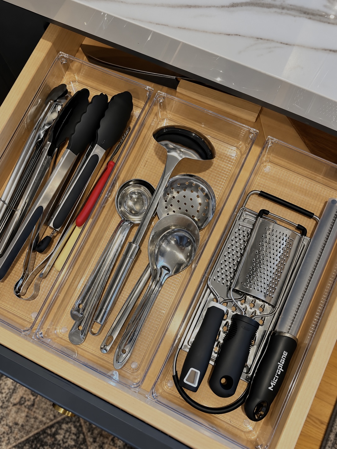 Open kitchen drawer with three clear bins holding tongs, ladles, and flat graters