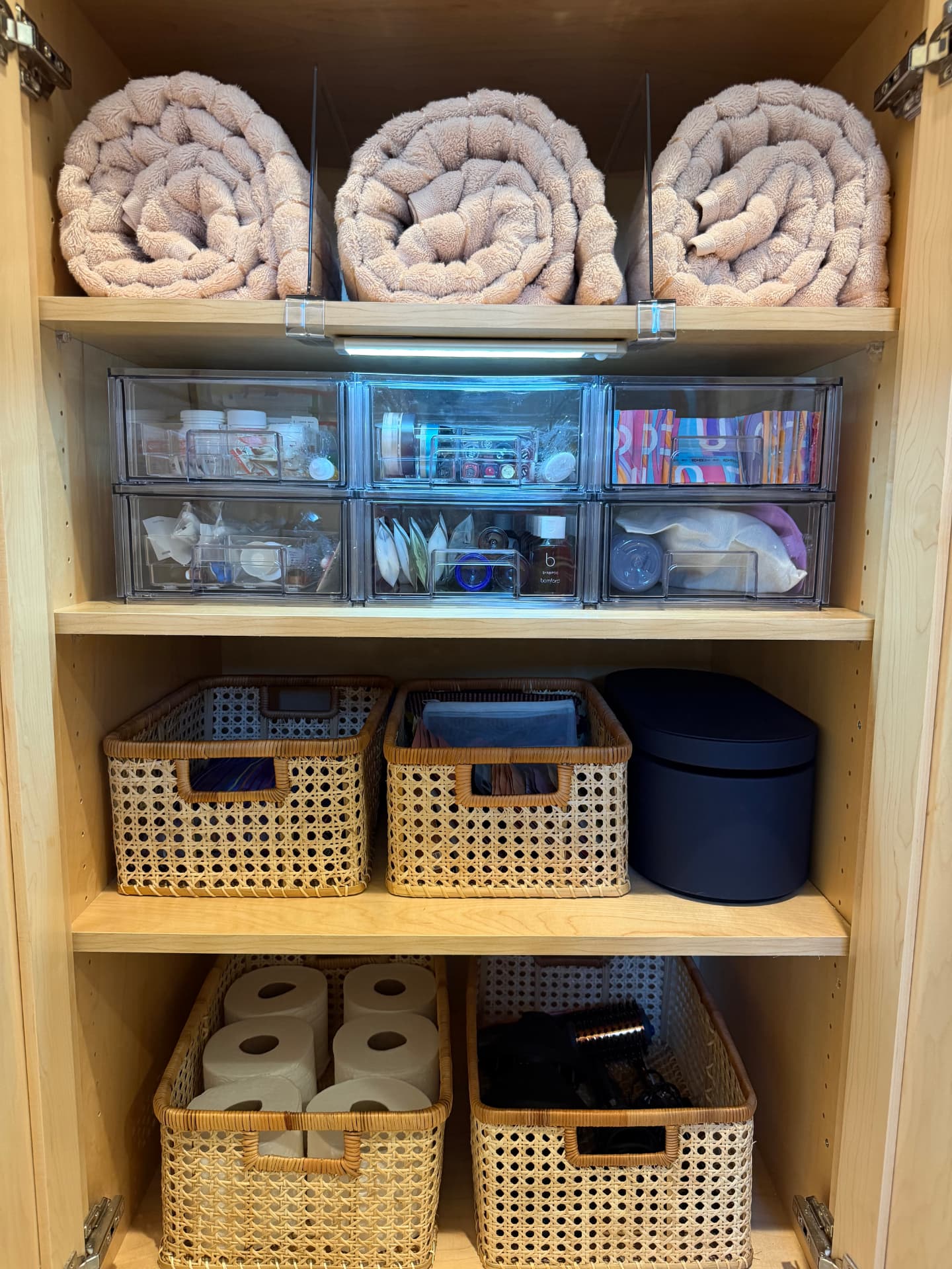 Lower shelves with rolled pink towels between clear dividers, six stacked clear drawers of toiletries, and cane baskets of toilet paper and hair tools