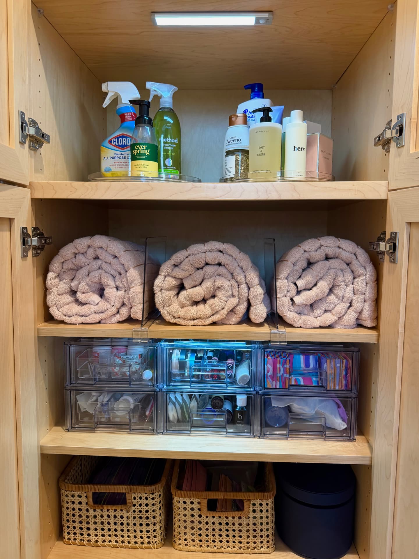 Organized bathroom storage closet with clear turntables of bath and cleaning products, rolled pink towels, clear drawers of toiletries, and woven baskets